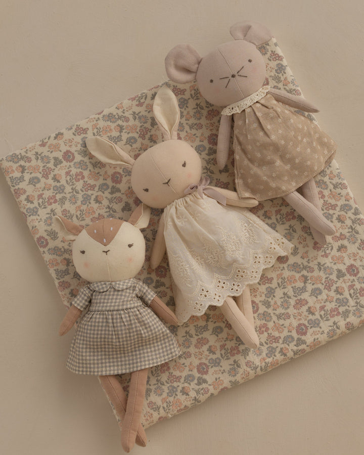 Three handmade plush bunny dolls in soft neutral tones, each wearing a different embroidered dress, arranged on a floral cushion against a warm beige background.