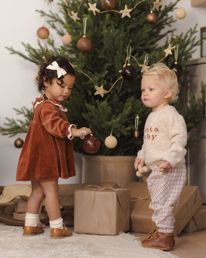 Two toddlers standing in front of a decorated Christmas tree with wrapped gifts on the floor. The child on the left wears a rust-brown velvet dress and brown shoes, while the child on the right wears a cream knit sweater, plaid pants, and brown boots.