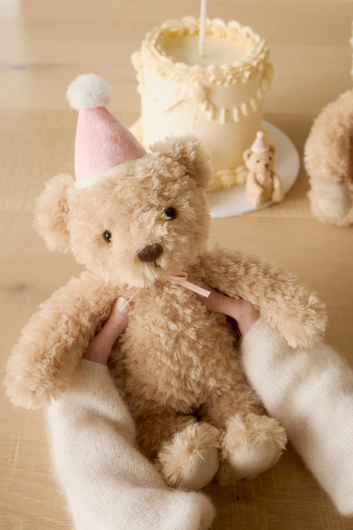Teddy bear wearing a pink party hat held by a person on a wooden surface with a blurred background.