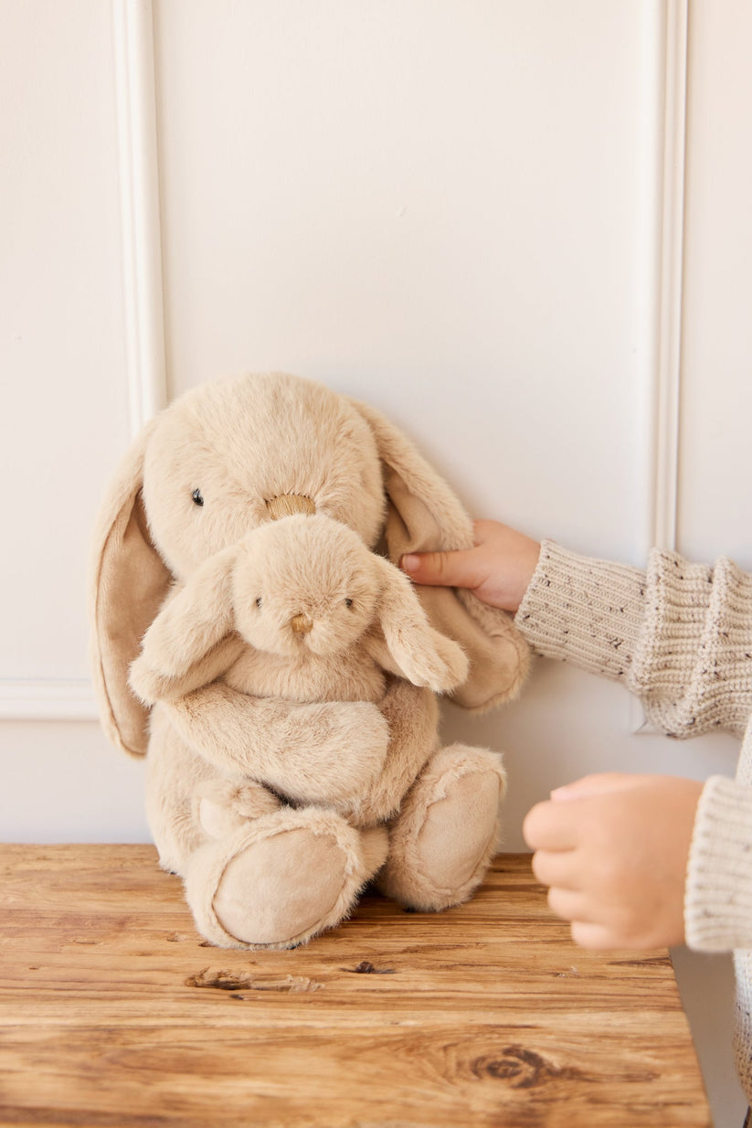 Two beige plush bunny toys on a wooden surface with a hand reaching towards them.