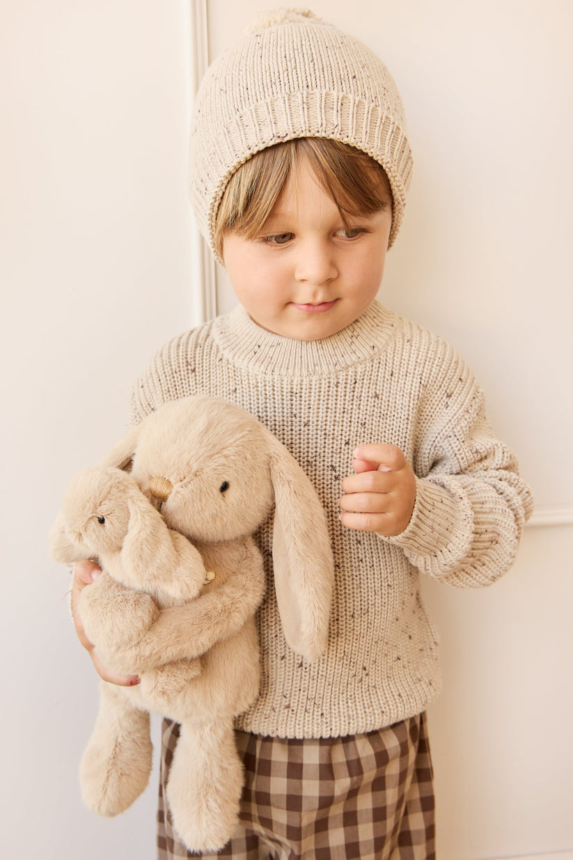 Child wearing a knit sweater and hat holding a plush bunny toy against a white wall.