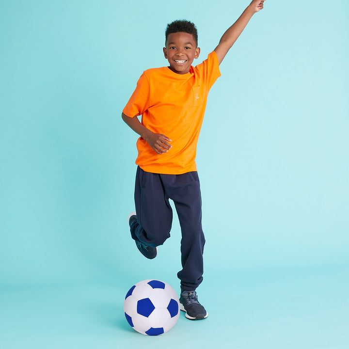 Child in orange shirt and dark pants playing with a soccer ball on a light blue background