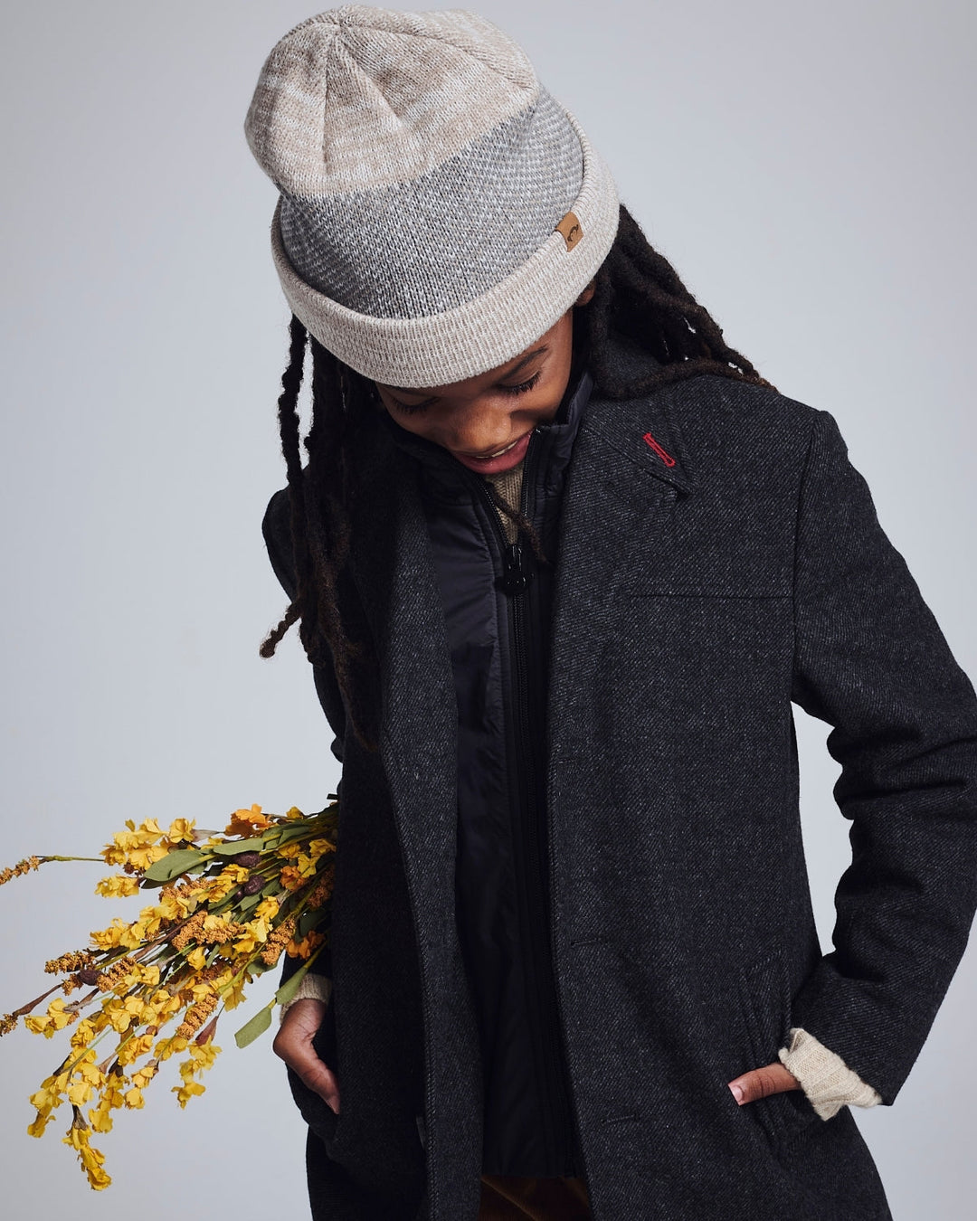 Person wearing a gray coat and patterned beanie holding yellow flowers against a light background