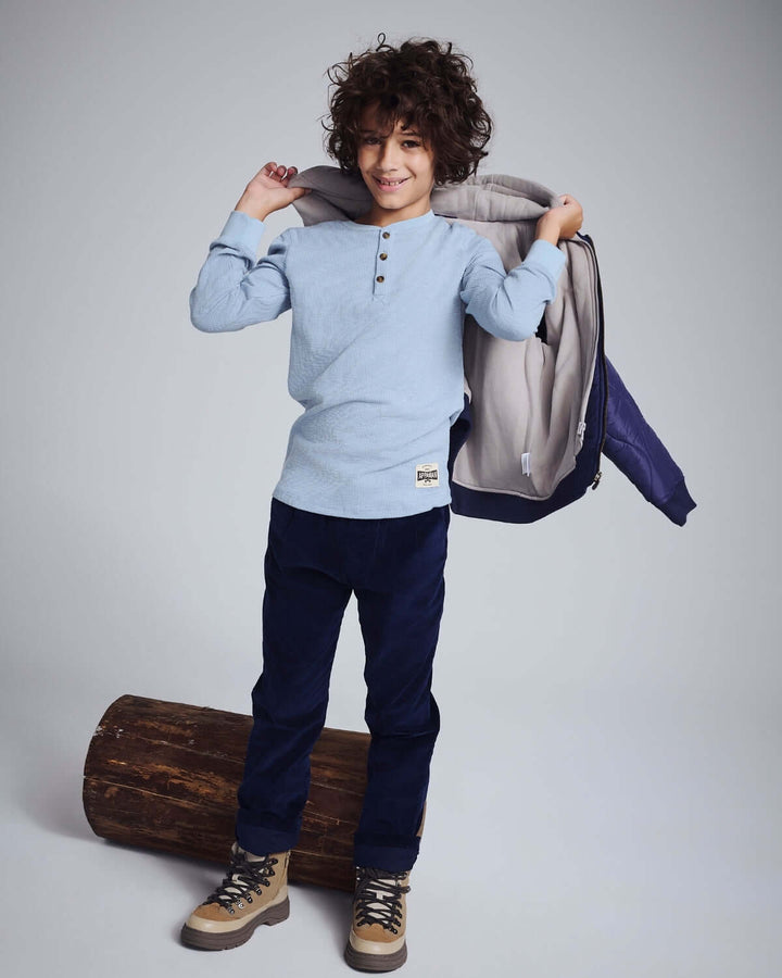 Child wearing a light blue sweater and dark pants, holding a gray jacket over their shoulder against a plain background.