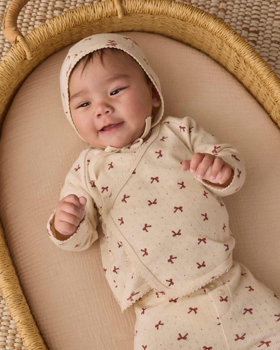 A smiling baby lying on their back inside a woven rattan bassinet, wearing a cream-colored knit romper and bonnet with a small brown bow pattern.