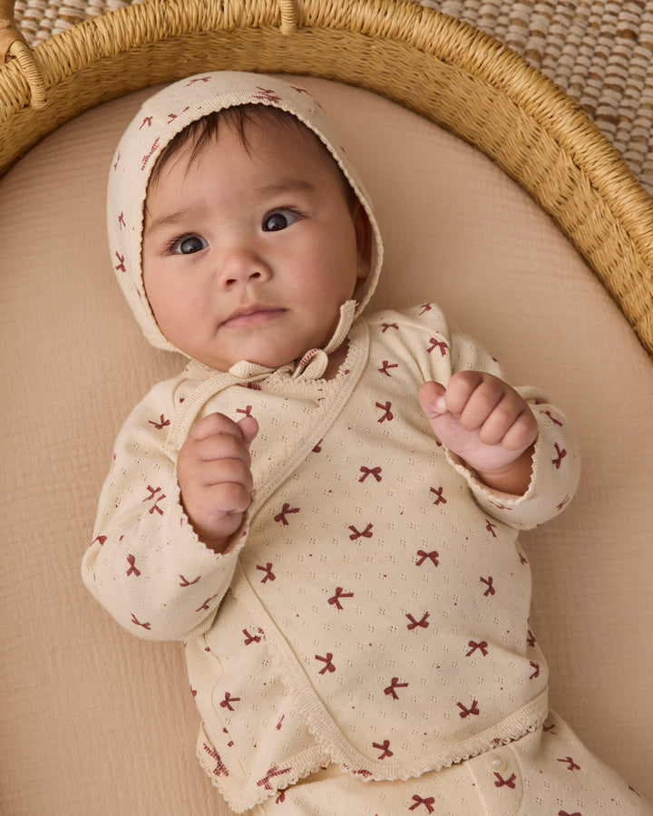 A baby lying on a beige cushion inside a woven bassinet, wearing a cream-colored hooded romper with a small red bow pattern.