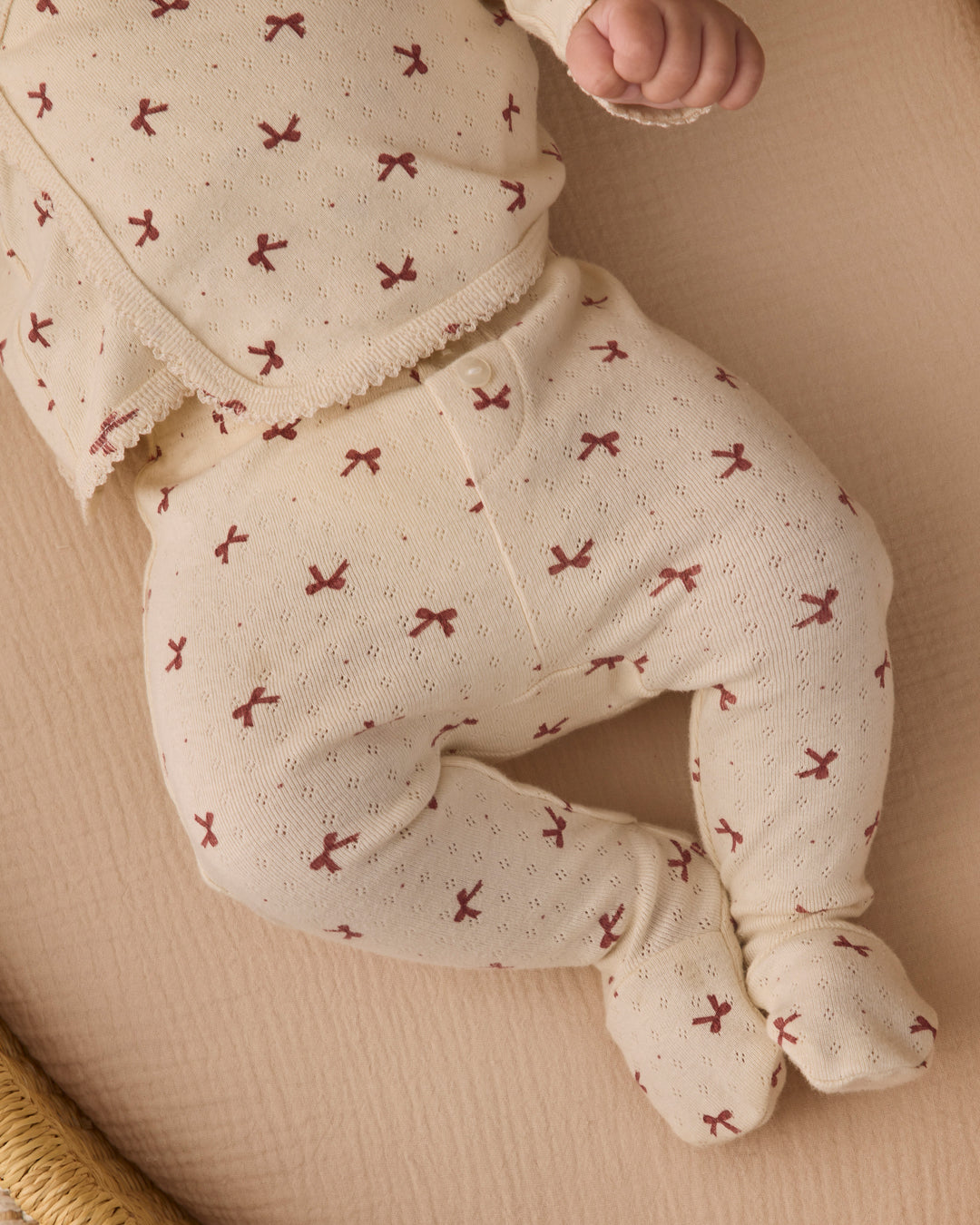 A baby wearing a cream-colored footed sleeper with a small red bow pattern, lying on a textured beige blanket.