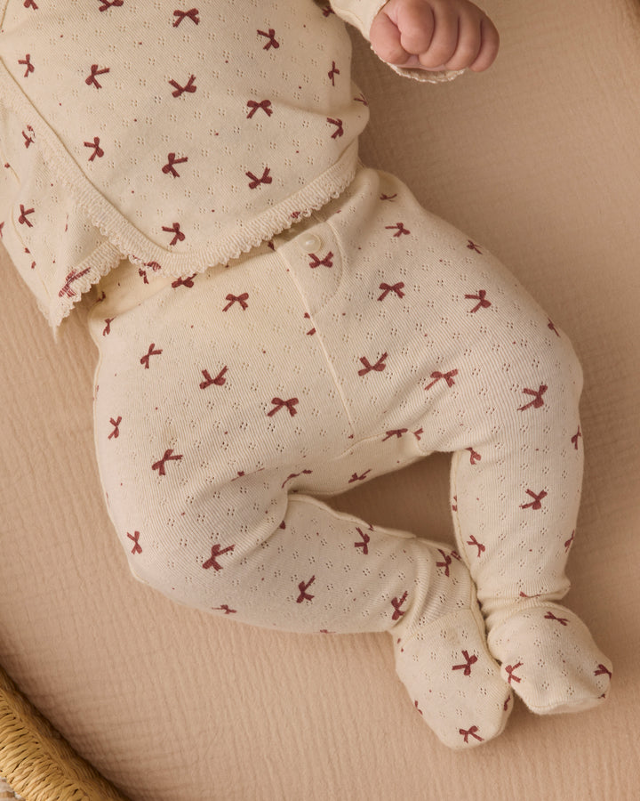 A baby wearing a cream-colored footed sleeper with a small red bow pattern, lying on a textured beige blanket.