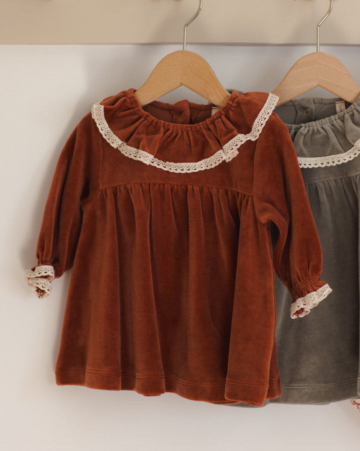 A rust-red long-sleeve children's dress with a cream lace ruffle collar and matching lace cuffs, hanging on a wooden hanger. A second gray dress is partially visible behind it on a hanger.