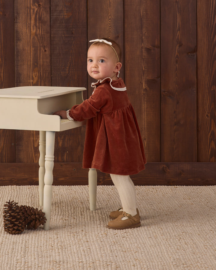 A toddler girl wearing a rust-red velvet dress with white trim, cream tights, and brown shoes stands next to a small cream-colored wooden table against a dark wood-paneled wall.