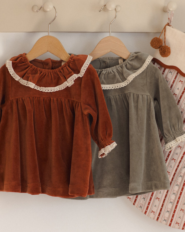 Two toddler girls' long-sleeve velvet tops hanging on wooden hangers on a wall. The front top is rust brown with a white lace ruffle collar and cuff, and the top behind it is muted gray-green with a similar white lace trim.