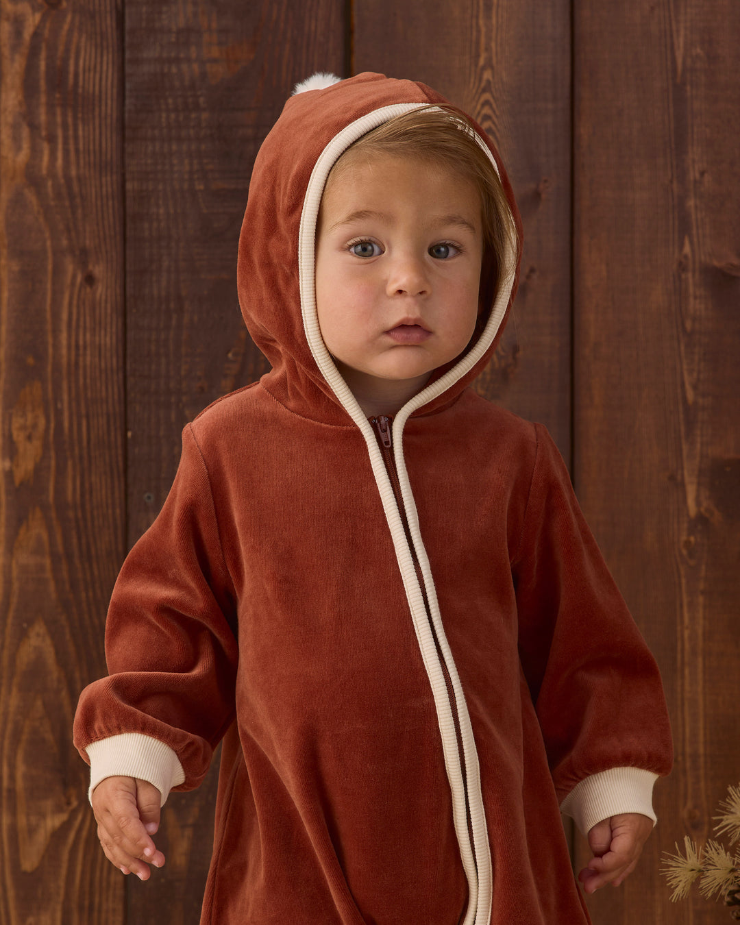 A toddler wearing a rust-red hooded fleece costume or onesie with cream trim, standing against a dark wood plank background.