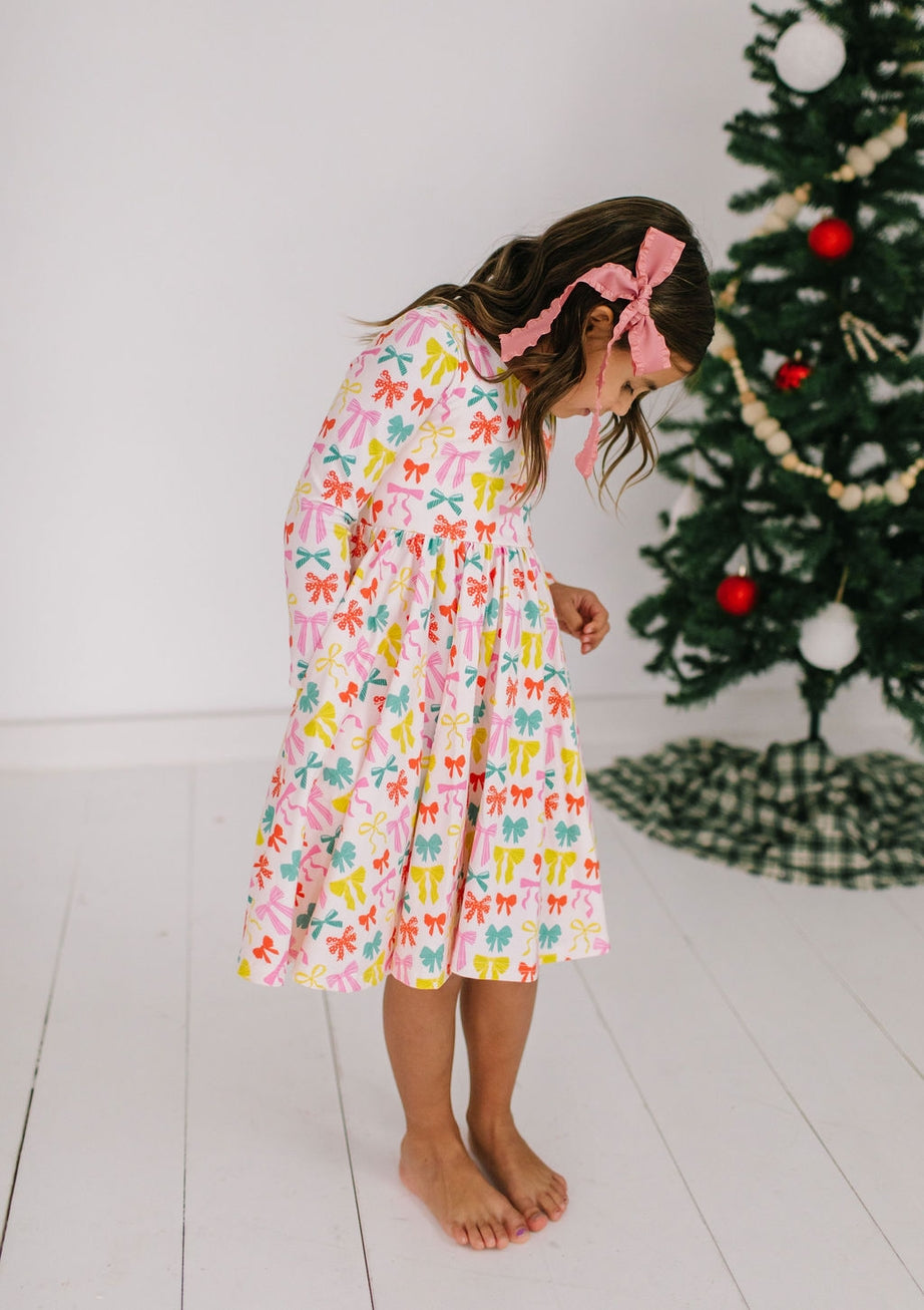 Young girl in a colorful dress with colorful bows all over it standing in front of a Christmas tree.