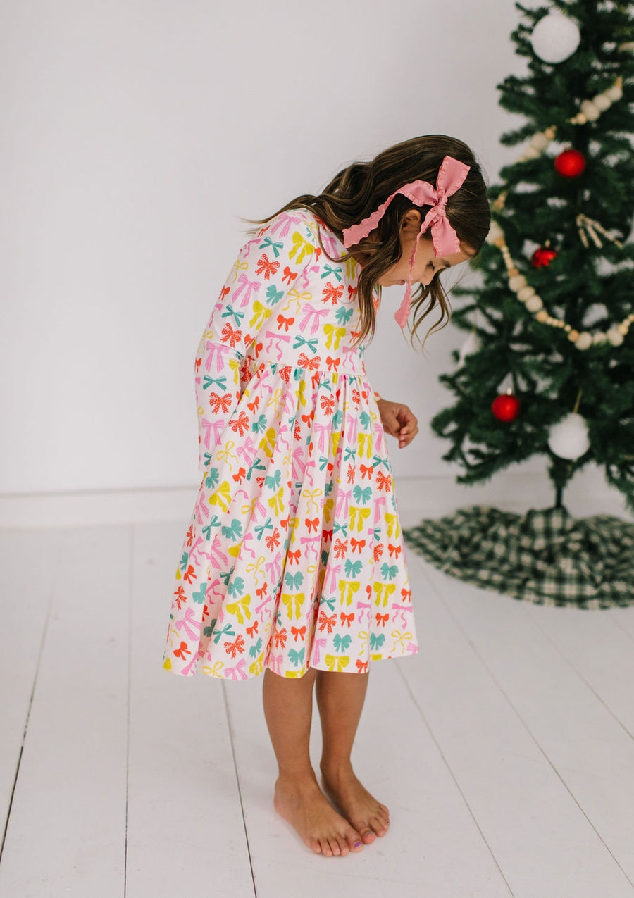 Young girl in a colorful dress with colorful bows all over it standing in front of a Christmas tree.