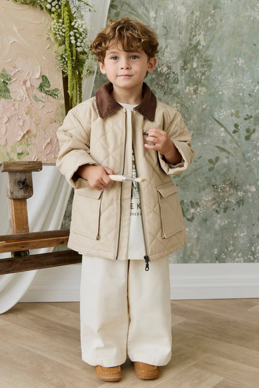 Child wearing a beige coat and pants standing in front of a floral wall.