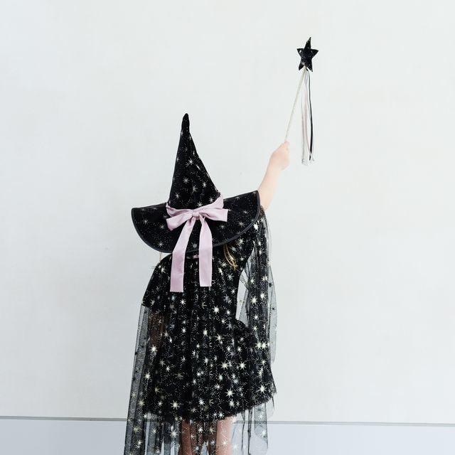 Person wearing a black sequin dress with a pink bow and witch hat, holding a wand with a star on a white background.