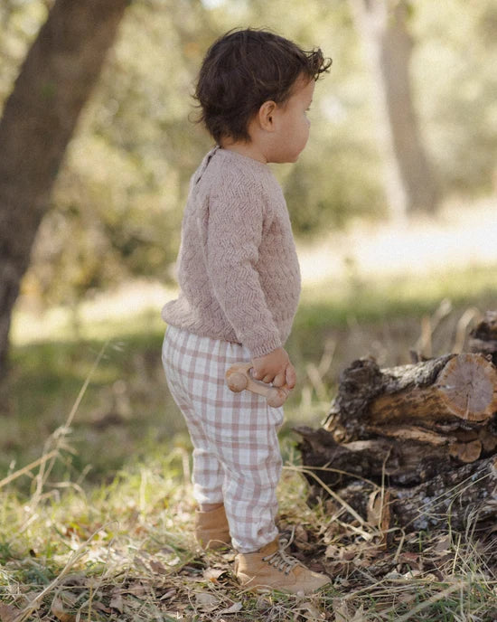 A toddler standing outdoors in a wooded setting, wearing a fuzzy light pink long-sleeve top, beige and white gingham pants, and tan boots.
