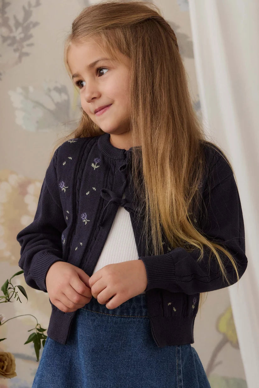 A young girl with long light brown hair wearing a navy blue knit cardigan with small embroidered details, layered over a white top and blue denim skirt, posing in an indoor setting with a floral wallpaper background.