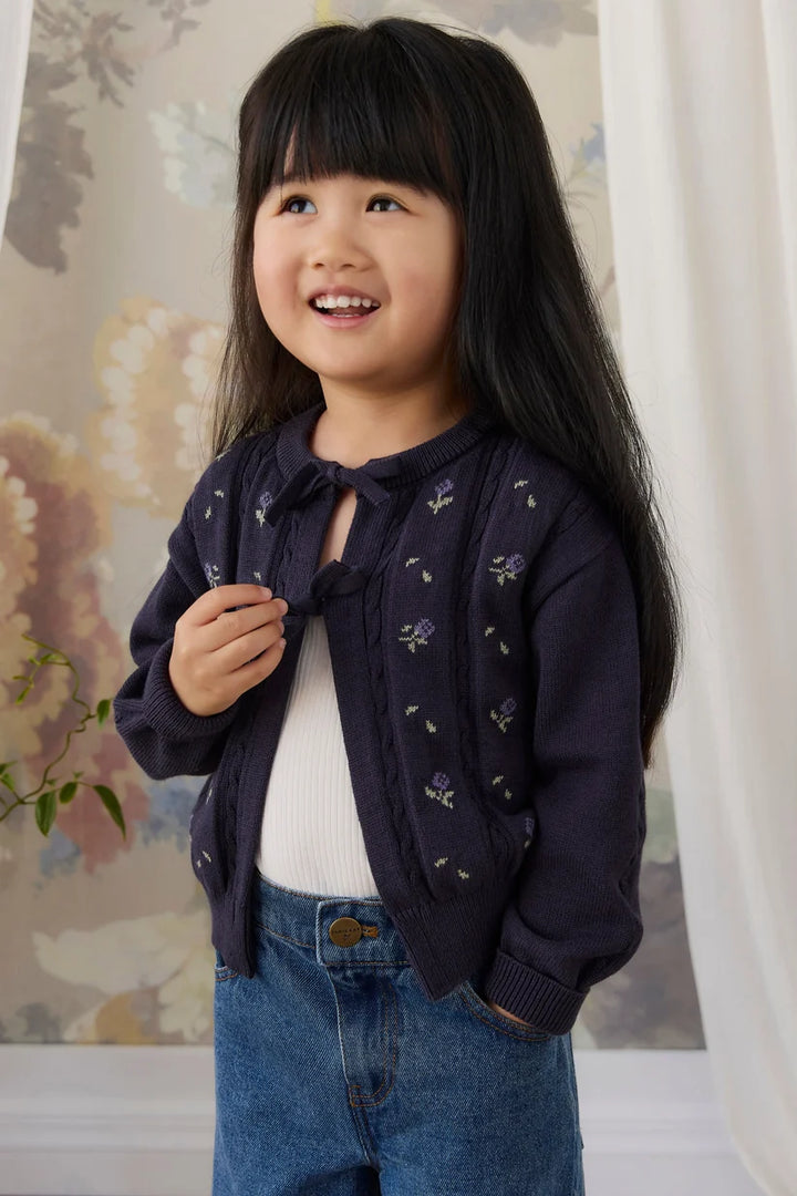 A young girl with long black hair smiling, wearing a dark navy blue knit cardigan with small floral embroidery over a white top and blue jeans, standing indoors against a floral wallpaper background.