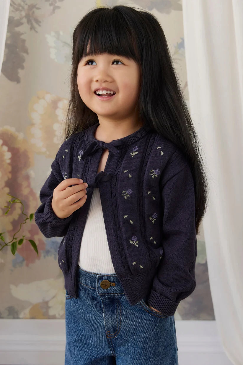 A young girl with long black hair smiling, wearing a dark navy blue knit cardigan with small floral embroidery over a white top and blue jeans, standing indoors against a floral wallpaper background.