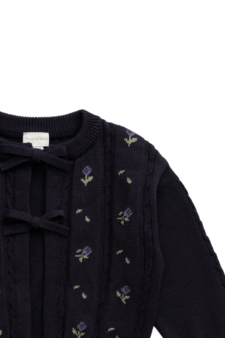 Close-up of a dark navy knit cardigan with purple and green floral patterns, featuring a tie-neck detail and a visible 'SWEETHEART' brand label.