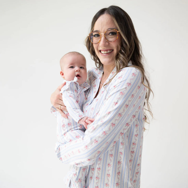 A woman wearing glasses and a white long-sleeve pajama shirt with pink floral and blue vertical stripes holds a baby dressed in a matching matching set against a plain white studio background.