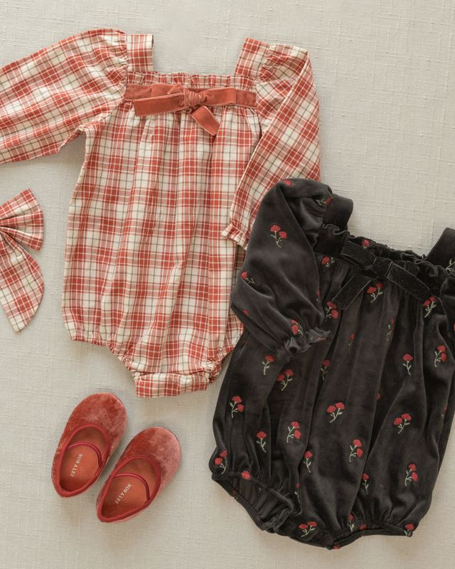 Two baby rompers, one red plaid and one black with red floral pattern, accompanied by red shoes on a light background.