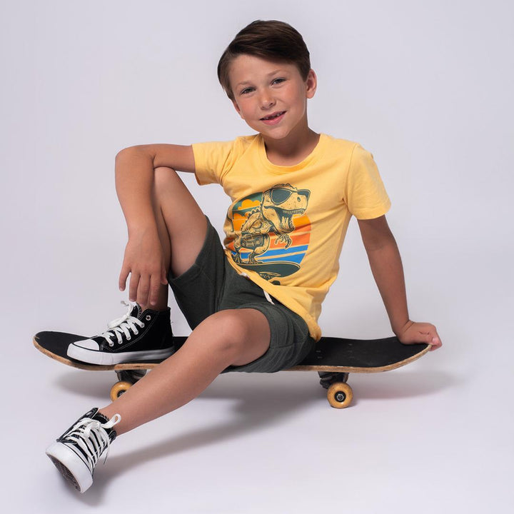 young boy wearing yellow tee with a dinosaur on it while sitting on a skateboard. 