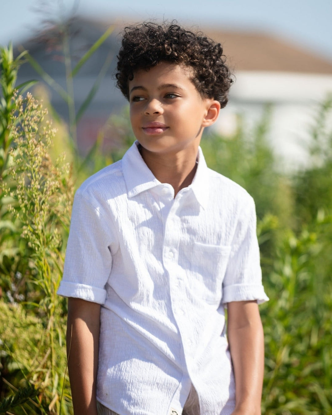 Child wearing a white shirt standing outdoors with greenery in the background