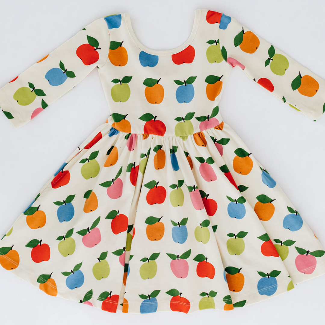 A long-sleeve kids dress with a white base and a colorful fruit print featuring red apples, orange pears, blue and green pears, and pink and red peaches. The dress has a gathered waist and a flared skirt, shown flat against a white background.