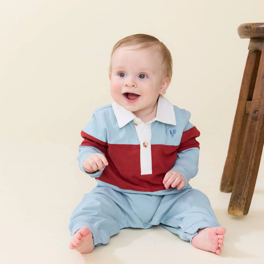 A baby sitting on a cream-colored floor wearing a light blue and red long-sleeve polo romper, with a wooden stool visible on the right side.
