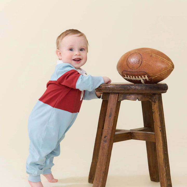 A baby wearing a color-blocked light blue and red one-piece outfit stands on a wooden stool next to a vintage brown leather football against a solid beige background.