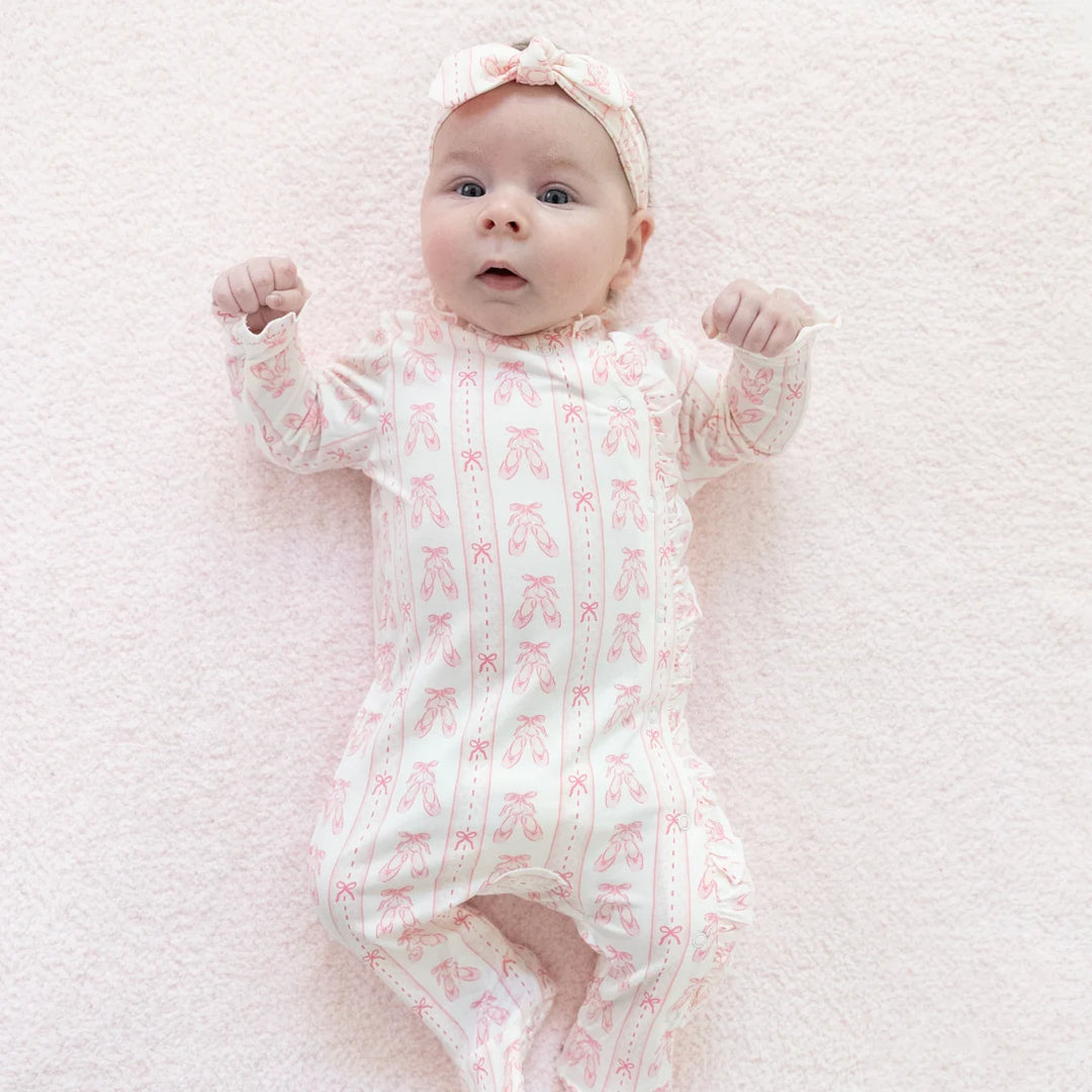 A baby lying on a soft pink blanket, wearing a long-sleeve footless onesie with a pink ballet shoe pattern and a matching pink bow headband.
