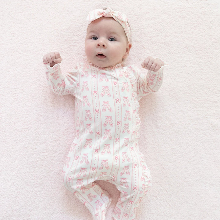 A baby lying on a soft pink blanket, wearing a long-sleeve footless onesie with a pink ballet shoe pattern and a matching pink bow headband.