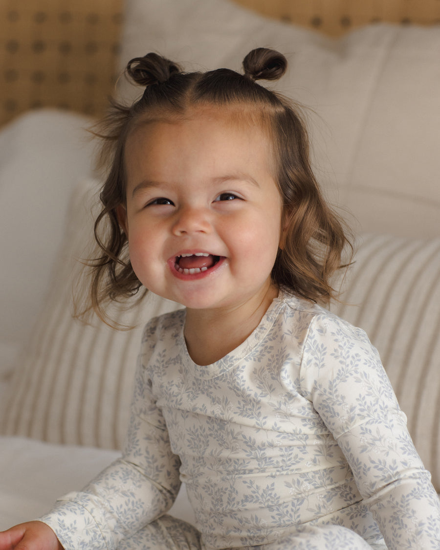 A smiling toddler with two buns in her hair wearing a long-sleeve cream pajama set with a delicate blue floral toile pattern, sitting on a bed with striped pillows.