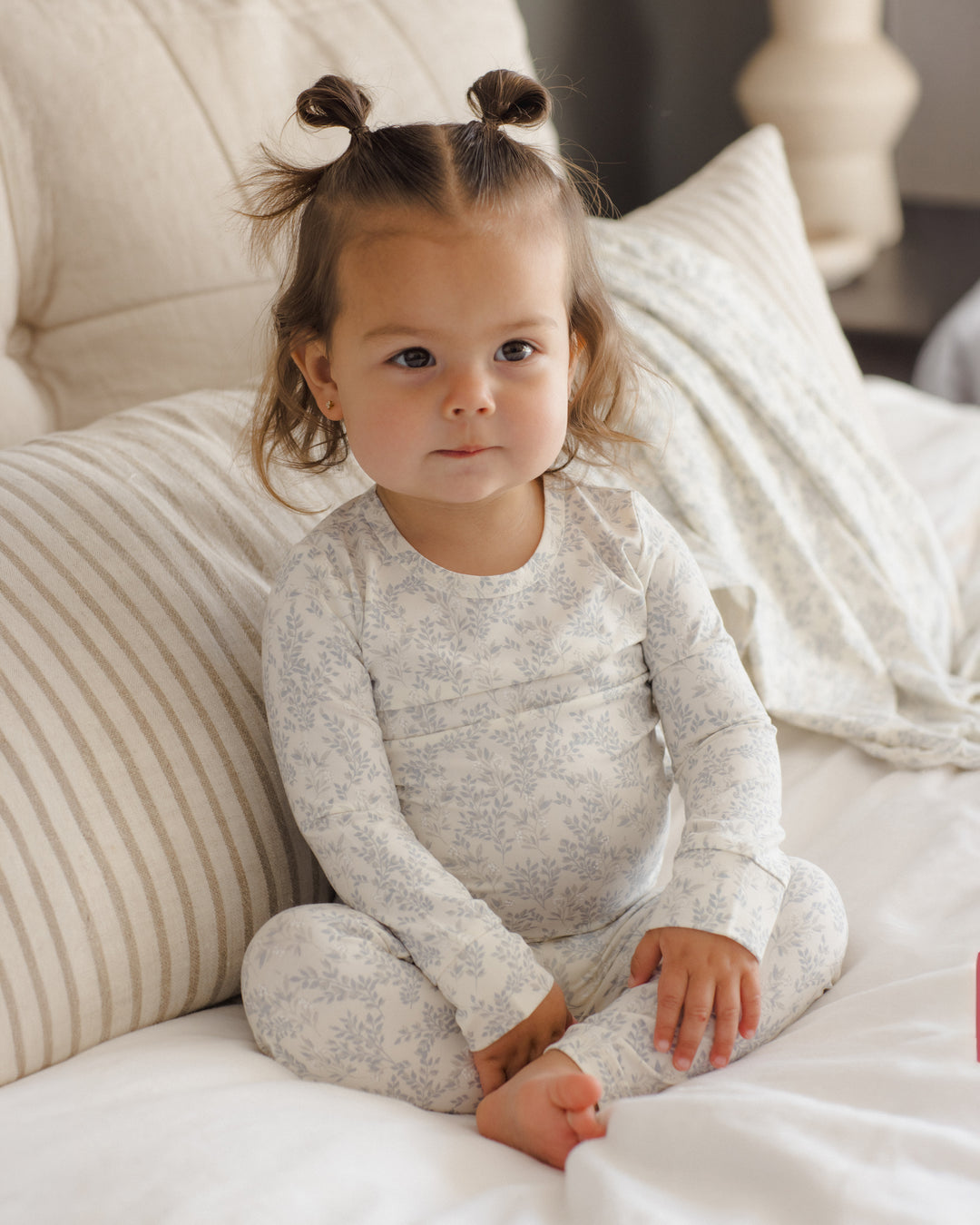 A toddler girl with two buns sits on a bed wearing a light blue and white floral print long-sleeve pajama set.