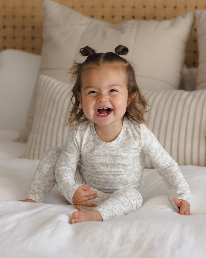 A young toddler girl with two buns sits smiling on a white bed, wearing a light blue and white patterned long-sleeve pajama set.
