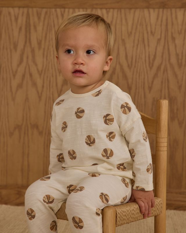Child wearing a white outfit with basketball patternoutfit sitting on a wooden chair against a wooden wall.