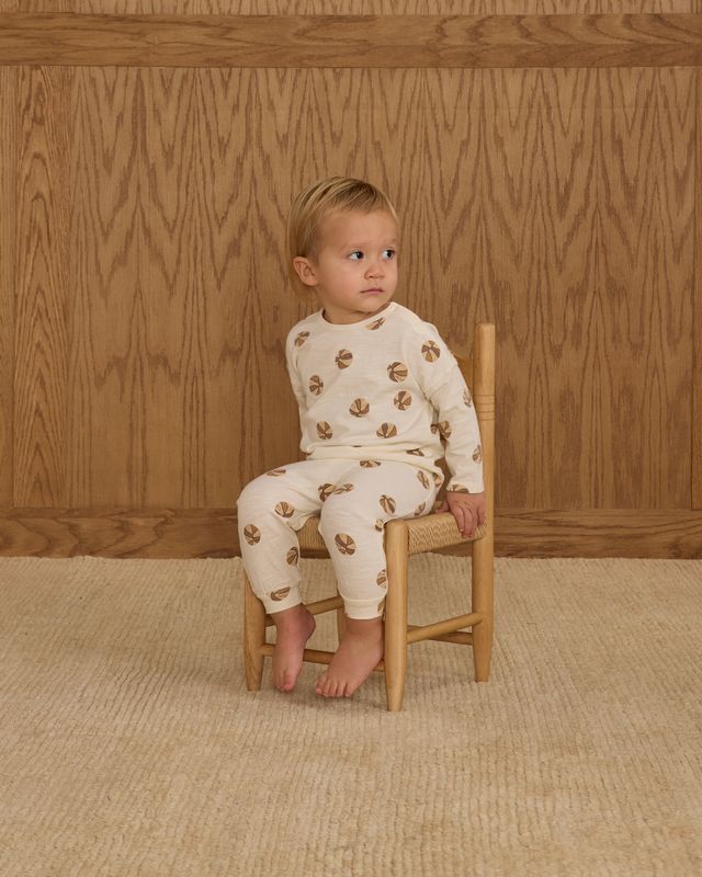 Child wearing a white outfit with basketball patterns sitting on a wooden chair against a wooden paneled wall.