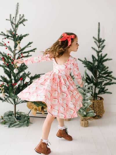 little girl twirling around in the holiday dress with santa mugs on it and christmas trees in the background. 