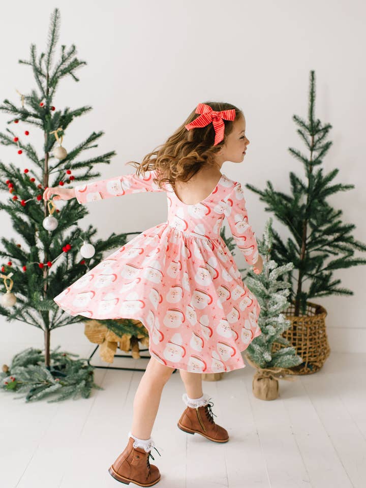 little girl twirling around in the holiday dress with santa mugs on it and christmas trees in the background. 