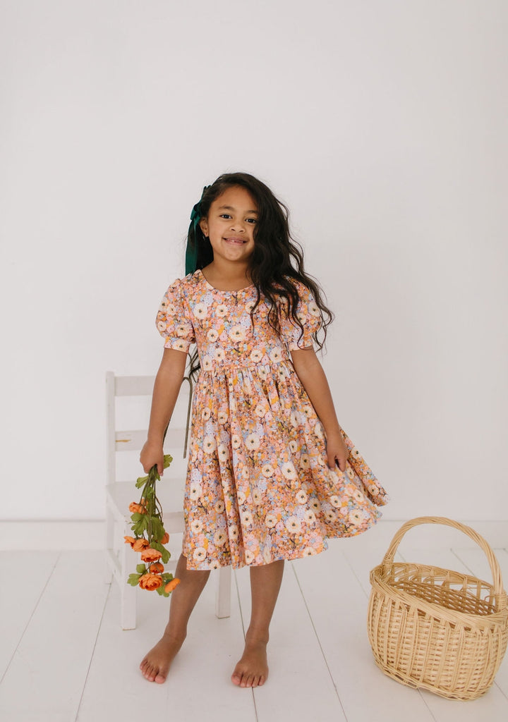 Young girl in a floral dress standing next to a basket on a white floor.