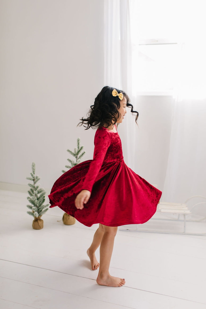 Child in a red velvet dress twirling in a bright room with small Christmas trees.