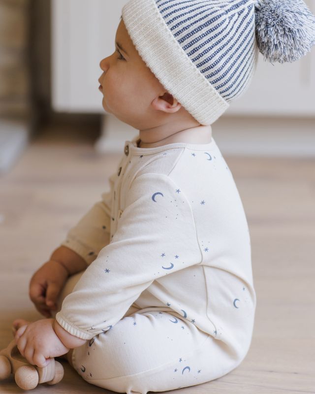 Baby wearing a white outfit with blue patterns and a matching hat, sitting on a wooden floor.
