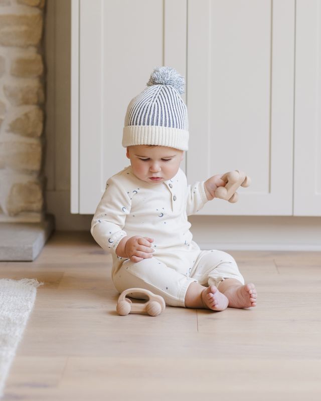 Baby sitting on a wooden floor wearing a white onesie and striped hat, with toys nearby.