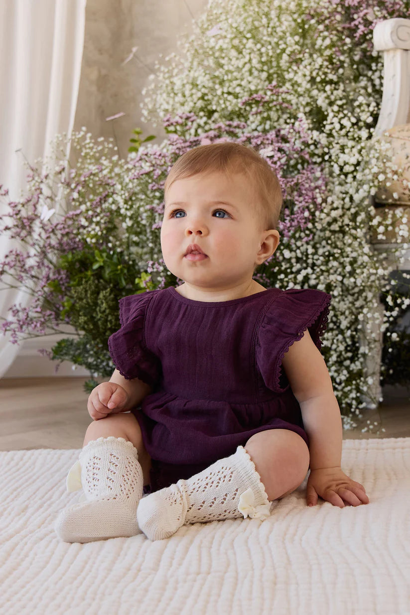 Baby in a purple outfit with white booties sitting on a blanket with flowers in the background
