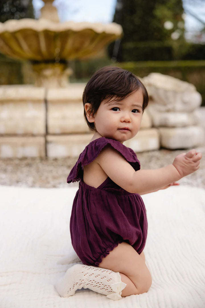 Baby in a purple romper sitting on a white surface with a fountain in the background