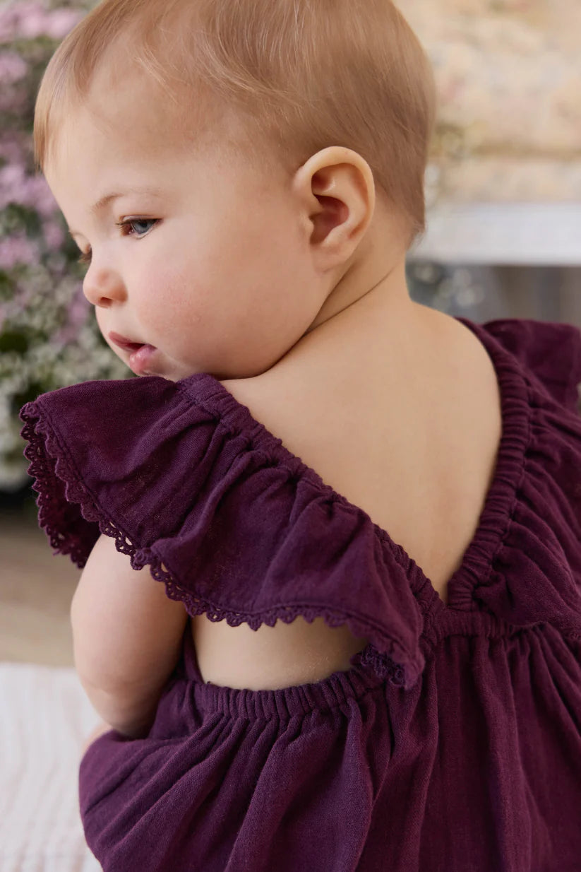 Baby wearing a purple romper with ruffled shoulders in a blurred indoor setting