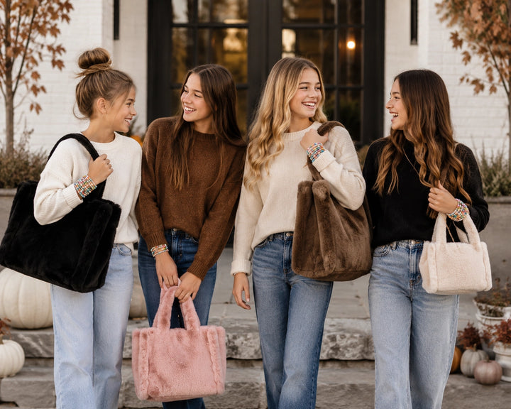 Four young women standing outdoors in front of a building, each carrying a large faux-fur tote bag in black, pink, cream, and light beige.