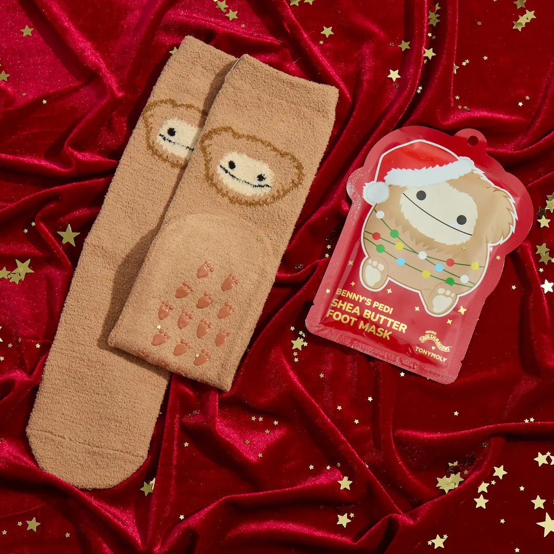 A pair of tan fleece socks with a sloth face and small heart details, displayed on a shiny red fabric backdrop with gold star confetti. A red hangtag for 'Pippi's Pedi Shea Butter Foot Mask' is also shown.